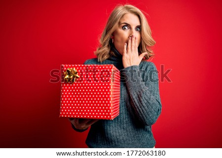 Middle age beautiful blonde woman holding birthday gift over isolated red background cover mouth with hand shocked with shame for mistake, expression of fear, scared in silence, secret concept
