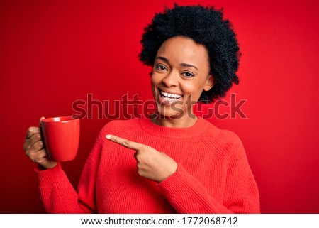 African American afro woman with curly hair drinking cup of coffee over red background very happy pointing with hand and finger