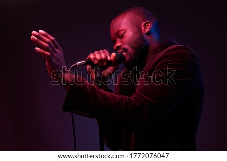 portrait of a dark-skinned handsome guy in dark jacket and t-shirt holds a microphone in his hands and emotionally sings in a dark studio with red and blue light Royalty-Free Stock Photo #1772076047