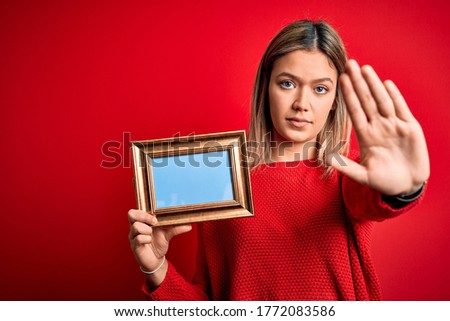 Young beautiful woman holding vintage frame standing over isolated red background with open hand doing stop sign with serious and confident expression, defense gesture