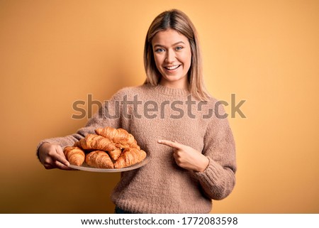 Young beautiful woman holding plate with croissants over isolated yellow background very happy pointing with hand and finger