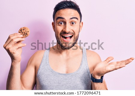 Young handsome man with beard wearing sleeveless t-shirt eating chocolate cookie celebrating victory with happy smile and winner expression with raised hands