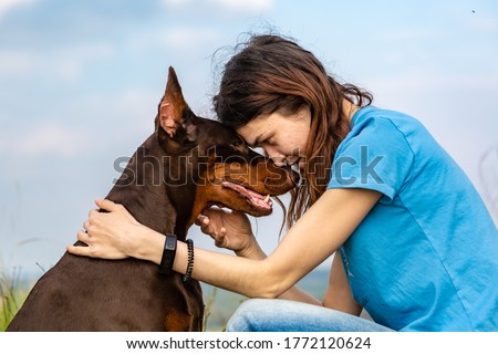 A girl in a blue T-shirt and a brown-and-tan doberman dobermann dog sit forehead to forehead against the sky. Horizontal orientation.  Royalty-Free Stock Photo #1772120624