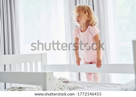 Standing on the bed. Cute little girl in casual clothes is indoors at home at daytime.