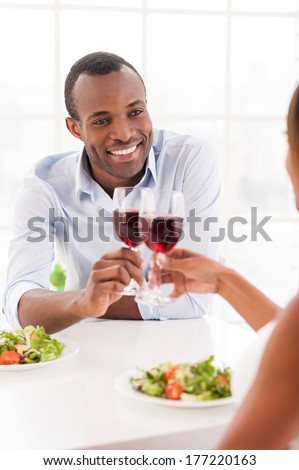 Having a dinner together. Cheerful young African couple sitting together at the table and holding wineglasses