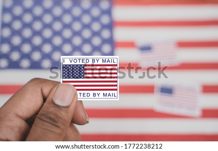 Holding I voted my mail sticker in hands with US flag as background - concept of voted through mail during election. Royalty-Free Stock Photo #1772238212