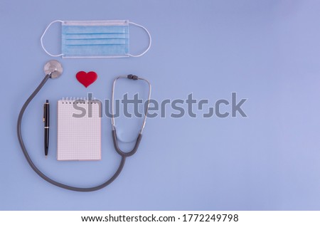 stethoscope, medical face mask and notepad with pen on blue background