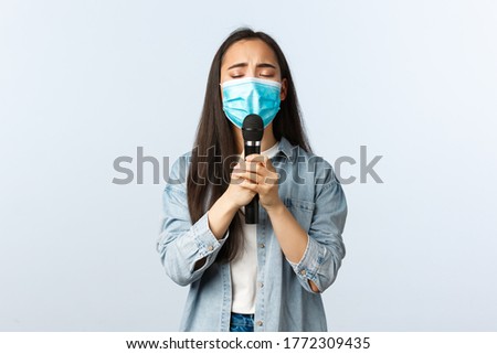Social distancing lifestyle, covid-19 pandemic and self-isolation leisure concept. Carefree passionate asian woman in medical mask singing with microphone, close eyes delighted