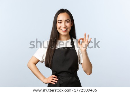 Coffee shop, small business and startup concept. Friendly charismatic asian barista show all good, okay sign and smiling. Cute waitress or cafe employee in black apron assure clients, make OK gesture