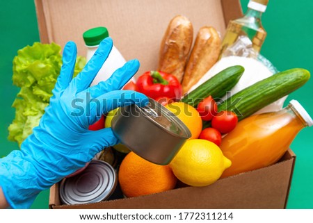 Big box with food supplies crisis food stock for quarantine isolation period on green.Vegetables, oil, herbs, bread isolated. Quarantine, food delivery,Donation.A gloved hand holds canned food.