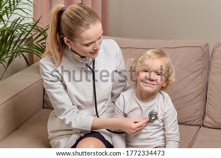 A female doctor sitting on a sofa listens to a child heart with a phonendoscope.