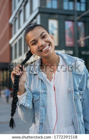 Authentic portrait of beautiful African American woman posing for pictures, smiling, walking on the street. Happy hipster girl with stylish hairstyle listening to music outdoors. Positive lifestyle