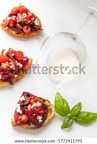 Fresh made Bruschetta with tomato, strawberry, caramelized onion, feta cheese and basil on a white surface with a wine glass. 
Ideal picture for magazine, blog, restaurant menu or site. 