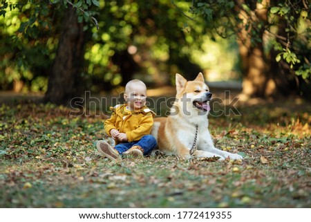 a beautiful little boy blond in an orange jacket sits with a red dog Akita inu on the ground in an autumn Park