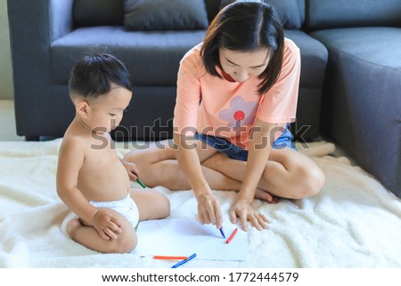 Asian mom teaching her cute baby boy to use crayons to draw on paper at home. Family and togetherness concept.