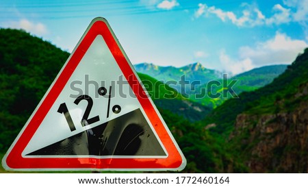 close up of road grade 12 per cent, road sign.
Red and white triangular warning road sign indicating a steep hill with a 12% slope ahead on a moorland road.
Mountain in background.
