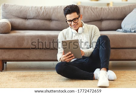 Young satisfied  male in casual clothes and wireless headphones surfing digital tablet while sitting on floor with legs crossed near soft couch in light modern living room
