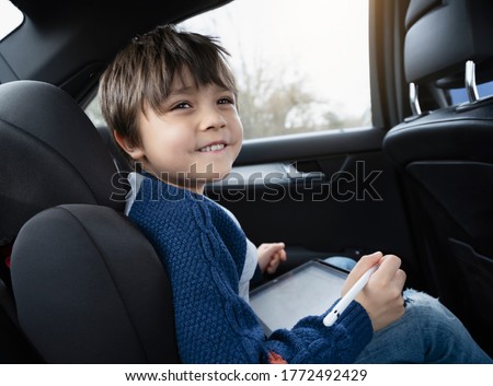 Happy young boy using a tablet computer while sitting in the back passenger seat of a car with a safety belt, Child boy drawing on smart pad,Portrait of toddler entertaining him self on a road trip. 
