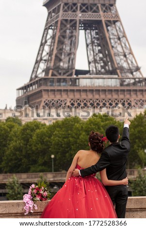 Soon to be married young couple is posing in front of the iconic Eiffel tower for their wedding photo album. Both bride and groom are formally dressed and cuddling. Groom is pointing the Eiffel tower.