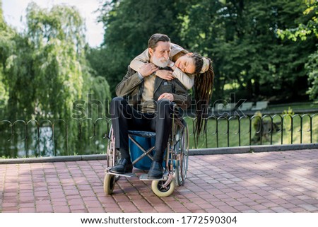 The old bearded man on a wheelchair and his pretty hipster granddaughter wirh dreadlock hair are walking in the park, hugging and looking each other, happy to spend time together