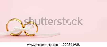 Elegant Wedding or Engagement banner background - pair of golden wedding rings and gentle white bow ribbon on gentle pink backdrop, copy space for text Royalty-Free Stock Photo #1772593988