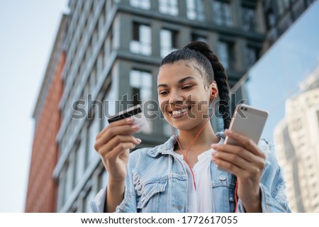 African American woman holding credit card, using smartphone for online shopping, mobile banking. Young freelancer receive payment, check balance. Hipster girl ordering food online, booking tickets