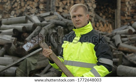 Portrait of lumberjack posing in green jacket with reflective stripes and holds big axe. Blonde man woodcutter standing and looking at camera. Sawn logs, firewood background. Sawmill, wood harvesting