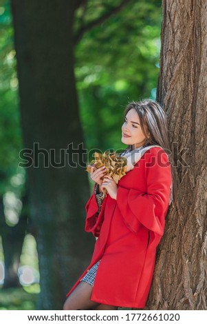 Portrait of young girl in the autumn weather in warm clothes near tree, holding a bundle of leaves.