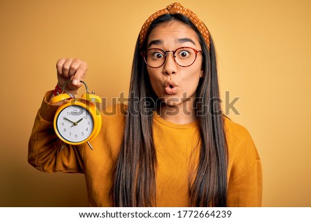 Young asian woman holding vintage alarm clock standing over isolated yellow background scared in shock with a surprise face, afraid and excited with fear expression