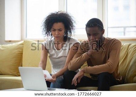 African spouses sitting on couch at home planning budget using laptop application check bills cheques feels troubled by overspending, American couple manage family expenses financial problems concept Royalty-Free Stock Photo #1772666027