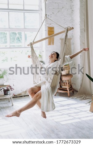 Vertical photo of carefree young adult female in bathrobe sitting on a hanging swing at home, enjoying lazy weekend morning, having fun and raised hands up, feeling free Royalty-Free Stock Photo #1772673329