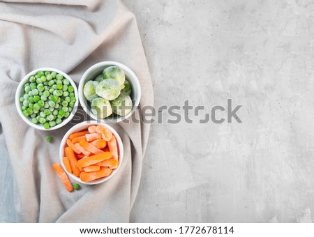 Frozen vegetables such as green peas, brussels sprouts and baby carrot in the white bowls on the concrete gray background with copy space, top view