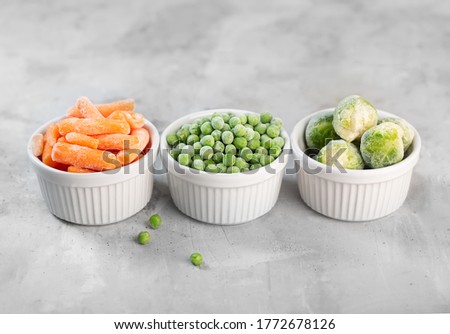 Frozen vegetables such as green peas, brussels sprouts and baby carrot in the white bowls on the concrete gray background