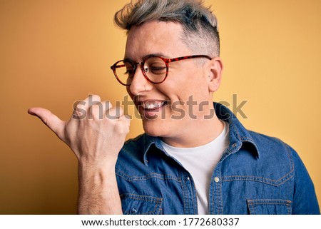 Close up of young handsome modern man wearing glasses and denim jacket over yellow background pointing and showing with thumb up to the side with happy face smiling