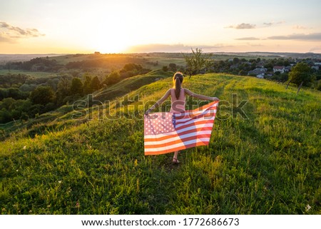 Back view of happy young woman posing with USA national flag outdoors at sunset. Positive girl celebrating United States independence day. International day of democracy concept.
