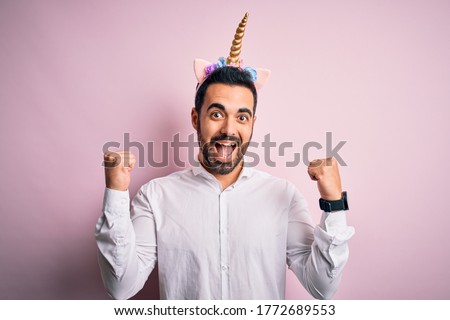 Young handsome man with beard wearing funny unicorn diadem over pink background celebrating surprised and amazed for success with arms raised and open eyes. Winner concept.