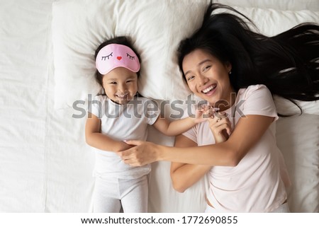 Above top view joyful smiling pretty biracial asian child daughter with sleeping mask relaxing on soft pillow with happy vietnamese mother, having fun together in cozy bed after waking up in morning.