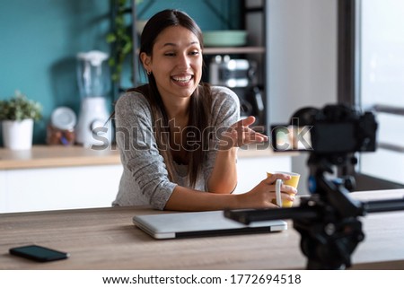 Shot of smiling young woman doing a video blog with the camera while holding a cup of coffee in the kitchen at home.