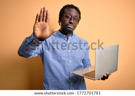 Young african american worker man working using laptop standing over yellow background with open hand doing stop sign with serious and confident expression, defense gesture