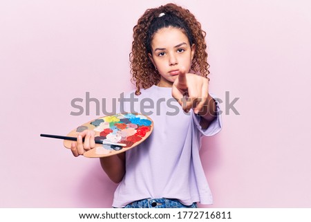 Beautiful kid girl with curly hair holding paintbrush and palette pointing with finger to the camera and to you, confident gesture looking serious 