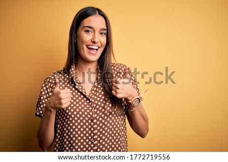 Young beautiful brunette woman wearing casual shirt over isolated yellow background success sign doing positive gesture with hand, thumbs up smiling and happy. Cheerful expression and winner gesture.