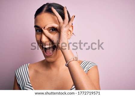 Young beautiful brunette woman wearing casual striped t.shirt over pink background with happy face smiling doing ok sign with hand on eye looking through fingers