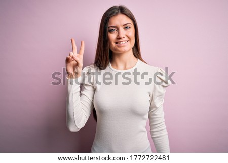 Young beautiful woman with blue eyes wearing casual white t-shirt over pink background smiling looking to the camera showing fingers doing victory sign. Number two.