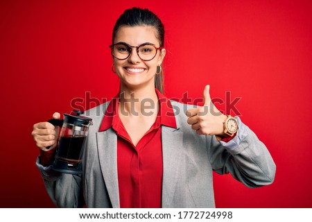 Young beautiful brunette woman doing coffe holding french coffeemaker over red background happy with big smile doing ok sign, thumb up with fingers, excellent sign