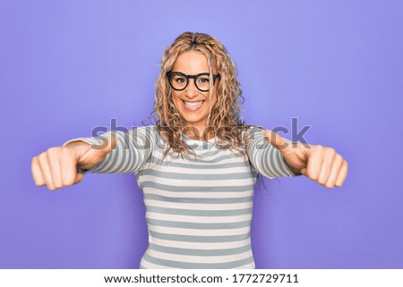 Beautiful blonde woman wearing casual striped t-shirt and glasses over purple background approving doing positive gesture with hand, thumbs up smiling and happy for success. Winner gesture.