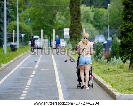 Rear view of a young blonde woman in shorts walking with a pram. Summer day in a city park. Walk with a small child in the fresh air. Copy space.
