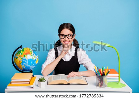 Photo minded girl university student sit table read textbook think thoughts touch hand chin look copyspace wear white blouse black overall ponytails uniform isolated blue color background