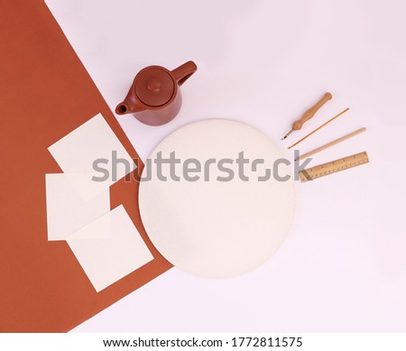 Blank fabric needle work frame in round shape with square paper mockups, punch and crochet needles, wooden ruler and pencil and a tea pot, top view creative concept image on white and tile red color