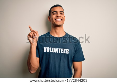 Young handsome african american man volunteering wearing t-shirt with volunteer message with a big smile on face, pointing with hand and finger to the side looking at the camera.