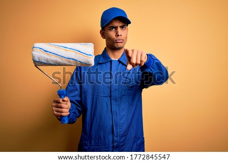 Young handsome african american painter man wearing uniform using painting roller pointing with finger to the camera and to you, hand sign, positive and confident gesture from the front
